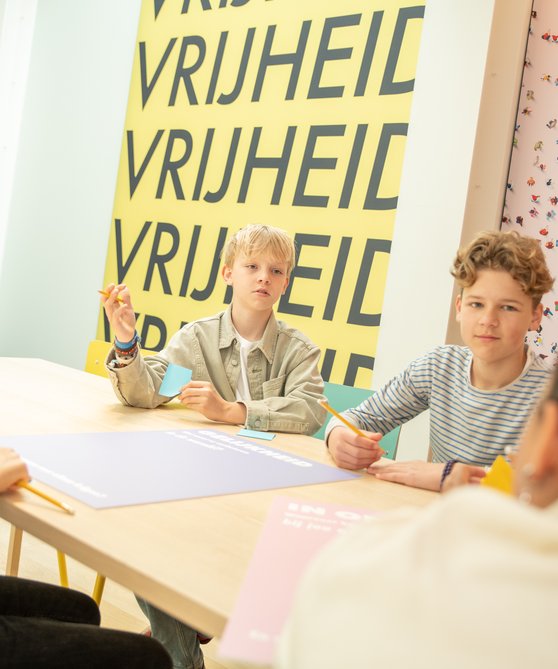 Er zitten kinderen aan een tafel in Hof van Nederland. Ze zijn aan het werk met het lesmateriaal.