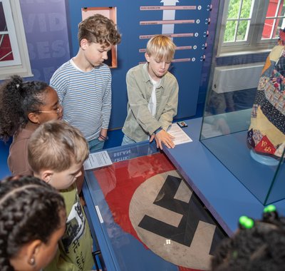 Een groepje kinderen staat voorovergebogen rond een vitrine met daarin een hakenkruisvlag in Hof van Nederland.