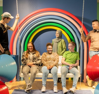 Drie jongeren zitten op de schommelbank, 2 staan aan de zijden. Achter hen een regenboog.