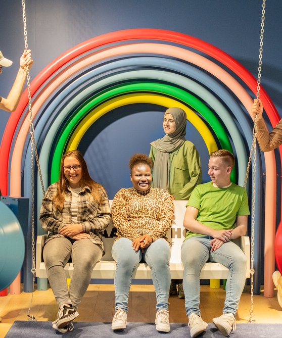 Drie jongeren zitten op de schommelbank, 2 staan aan de zijden. Achter hen een regenboog.