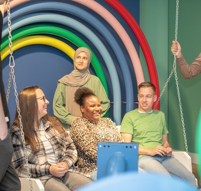 Een groep jonge mensen zit op de schommel in Hof van Nederland. Achter hen een regenboog op de wand.