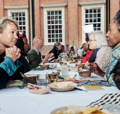 Mensen zijn met elkaar in gesprek tijdens een Keti Koti Tafel. Ze zitten aan een lange tafel waar eten en drinken op staat.