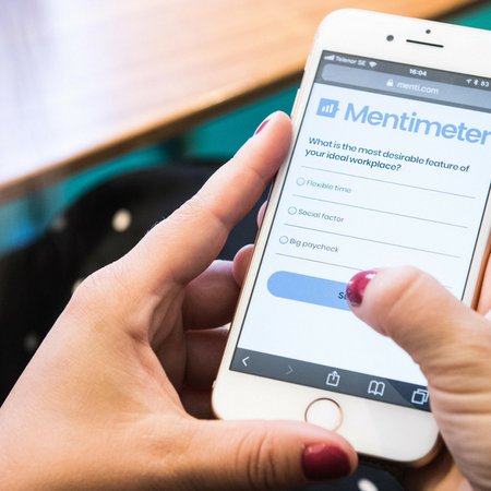 Twee handen houden mobiel vast. Er staat Mentimeter op het scherm.