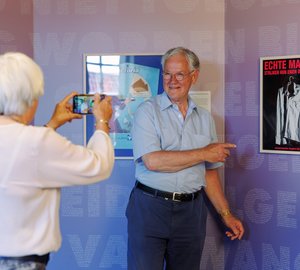 Een dame maakt een foto van een heer die naar een poster wijst met de tekst: echte mannen strijken hun eigen overhemd.