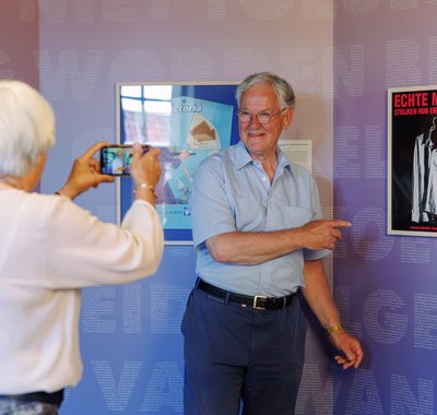 Een dame maakt een foto van een heer die naar een poster wijst met de tekst: echte mannen strijken hun eigen overhemd.