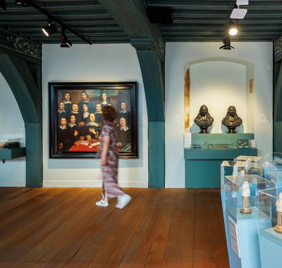 Een vrouw in lange jurk loopt door een museumzaal met schilderijen en voorwerpen in vitrines.