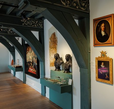 Een wand met schilderijen van personen in het museum. Er staan voorwerpen en bustes van de De Witten.