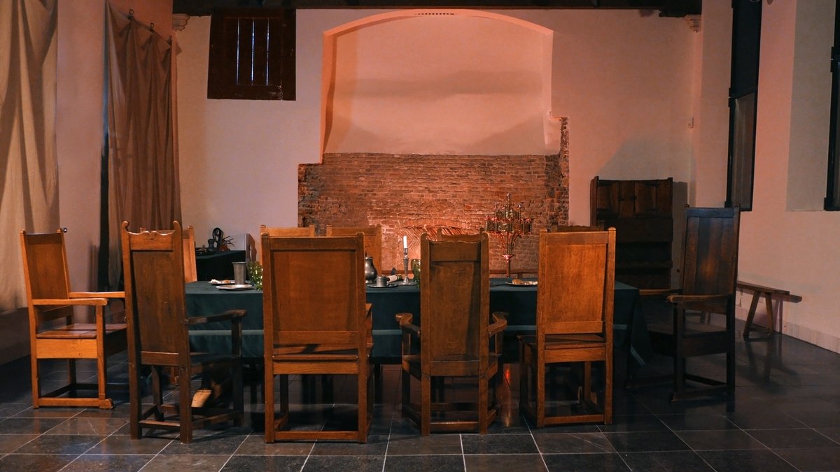 Zaal zonder mensen. Er staat een tafel met stoelen er om heen. Er brandt een kaars op tafel.