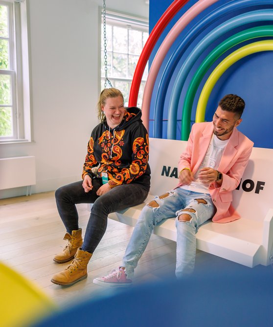 Een jonge man en vrouw zitten op een schommelbank in het museum. Ze lachen. Achter hen een regenboog aan de wand.