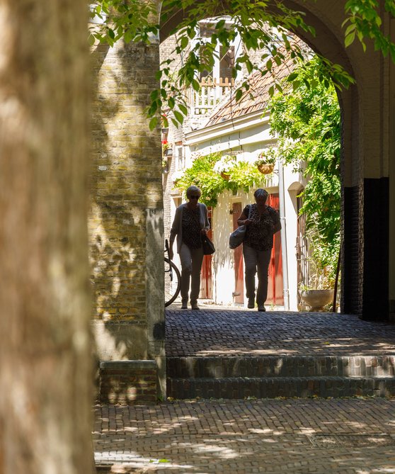 Twee dames komen aangelopen vanuit het steegje naar Hof van Nederland. Ze dragen schoudertassen.